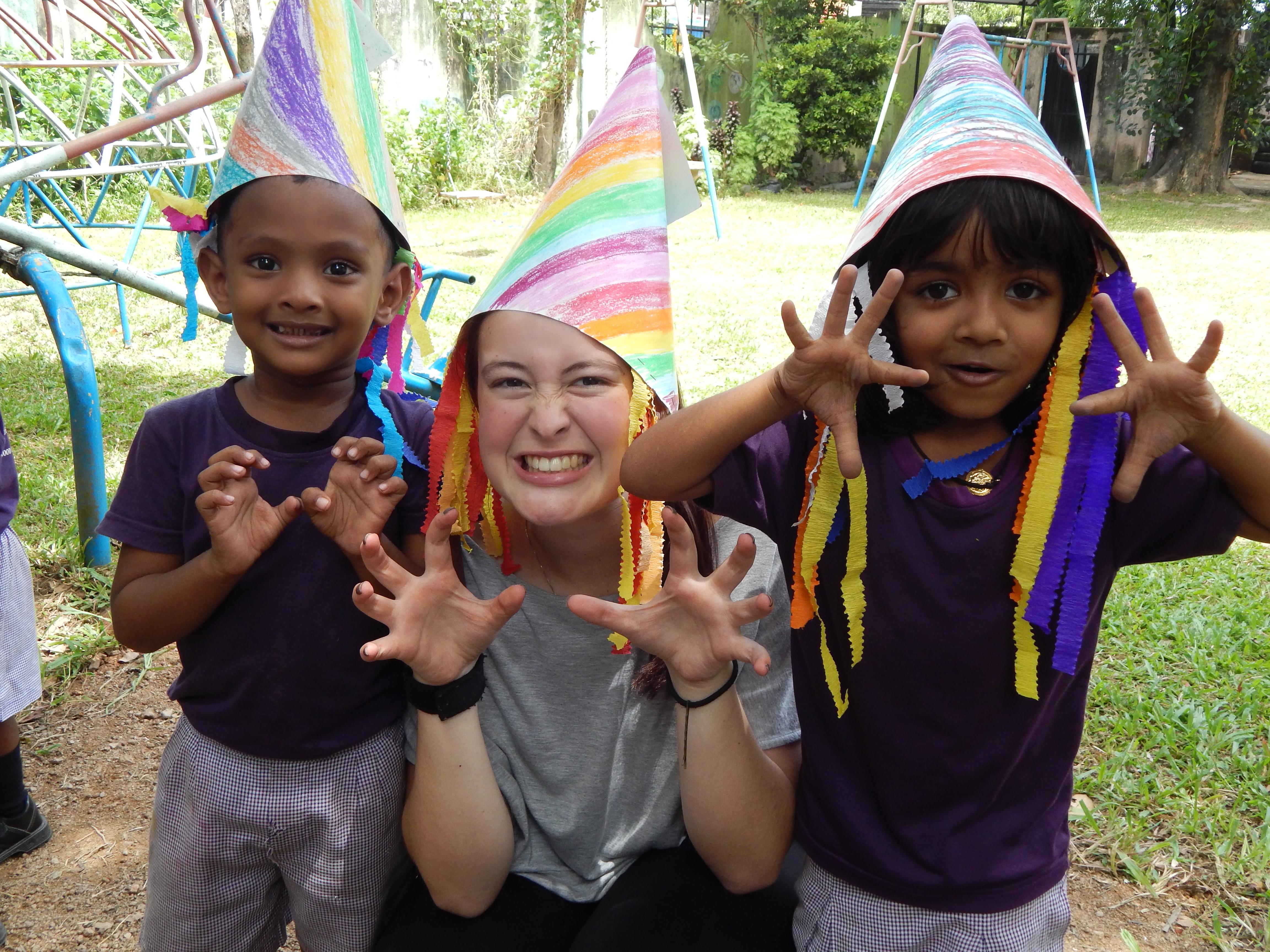 Projects Abroad Childcare volunteer with children in Sri Lanka plays dress up with two children outside a kindergarten. Female Childcare volunteer and children wear hats they have made during a fun activity on a Care placement in Sri Lanka.